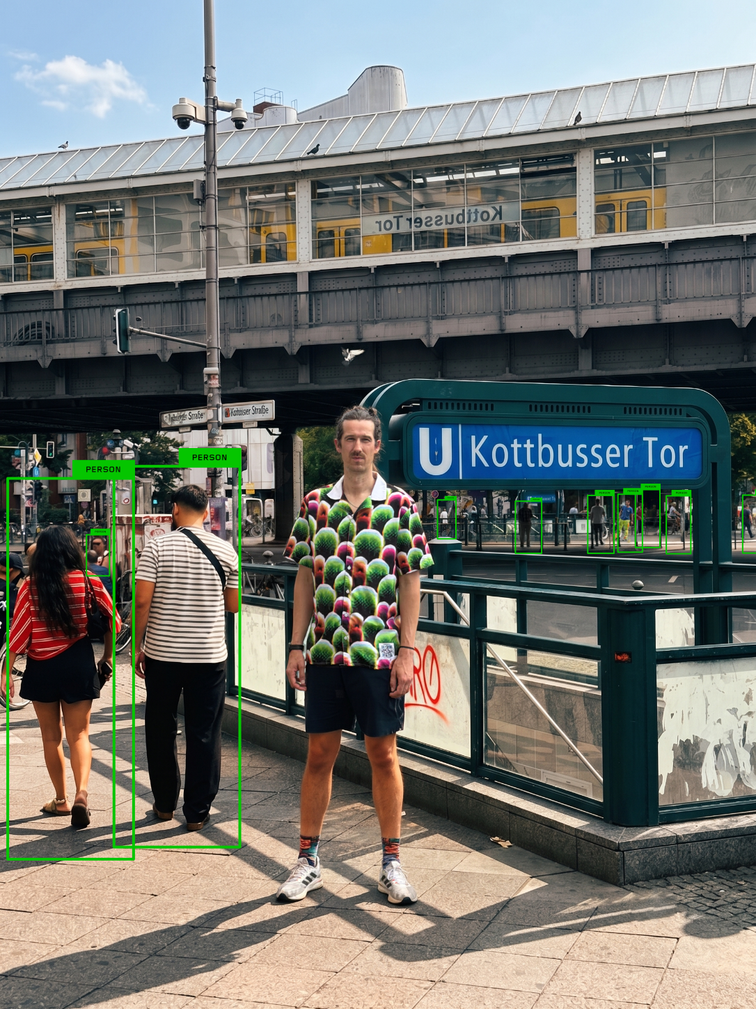 Homme portant la chemise Digital Camouflage à Kottbusser Tor à Berlin, entouré de piétons détectés par des cadres verts d'IA, lui n'étant repéré par aucun cadre.