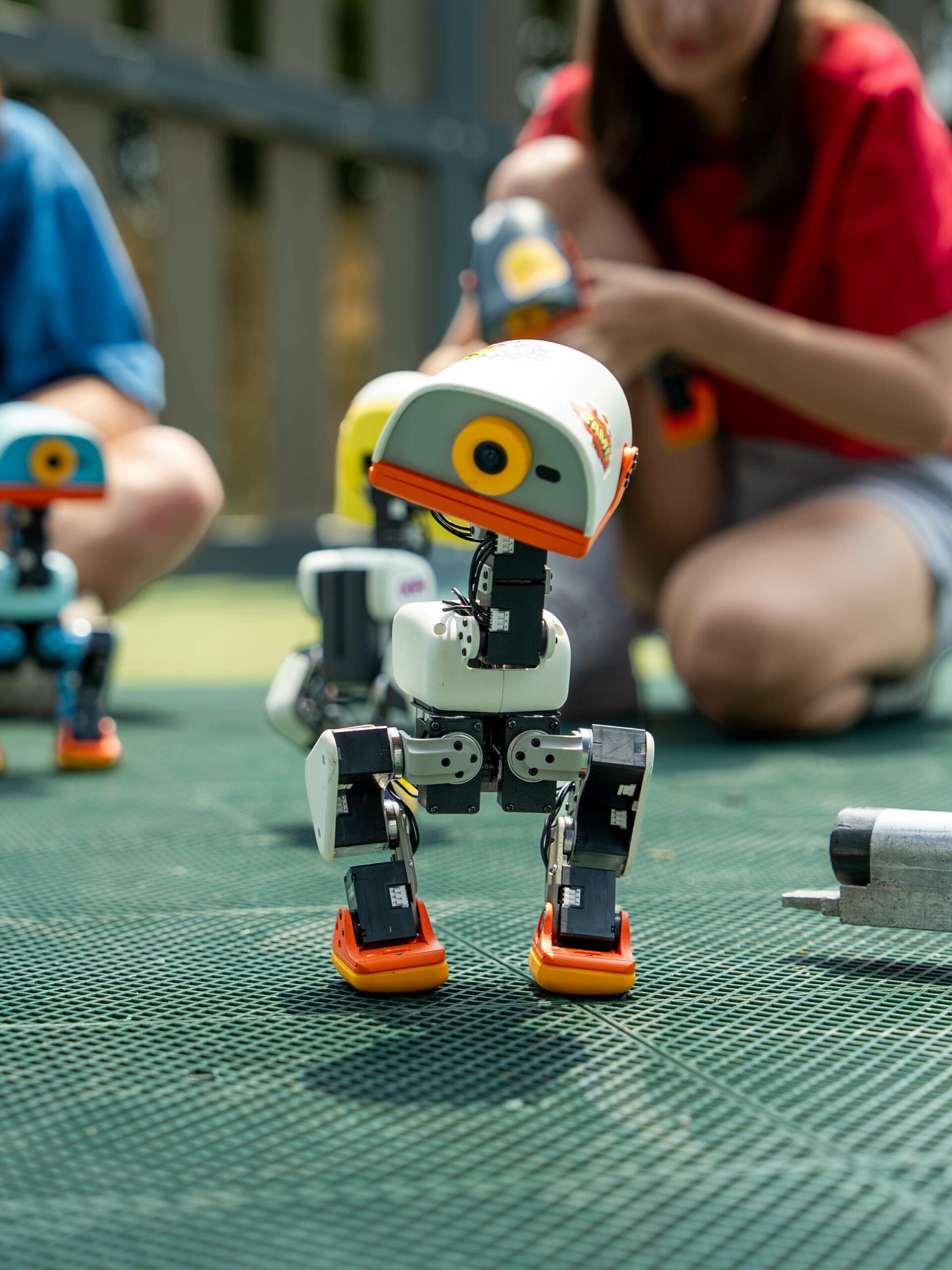 Robot Microduck bipède orange et blanc en extérieur, entouré d'autres exemplaires colorés, avec des personnes accroupies en arrière-plan qui les manipulent