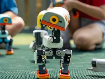 Robot Microduck bipède orange et blanc en extérieur, entouré d'autres exemplaires colorés, avec des personnes accroupies en arrière-plan qui les manipulent